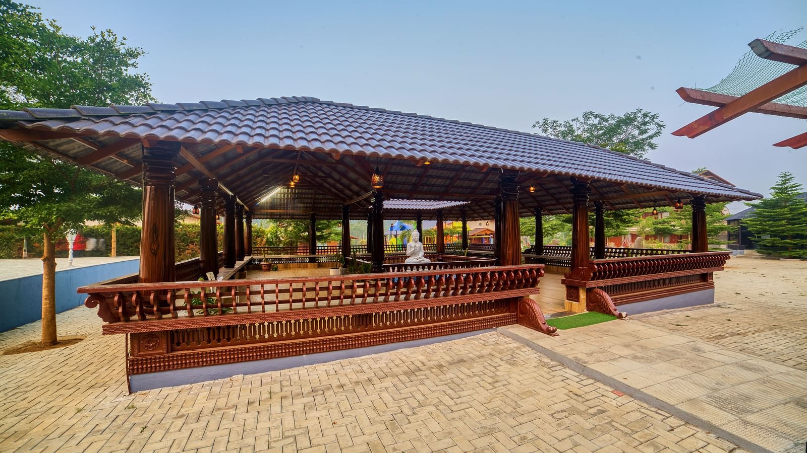 A large, open-sided wooden seating with a dark tiled roof, set on a paved area under a clear blue sky - Vintage Trails by Ilara