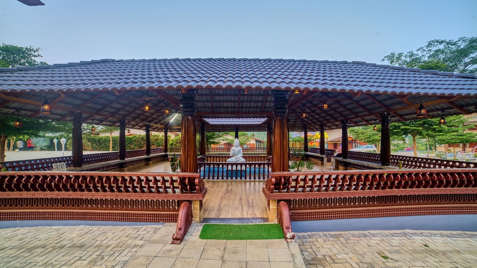 An open-sided wooden pavilion with a dark tiled roof and built-in seating, situated on a paved area, under a clear blue sky. - Vintage Trails by Ilara