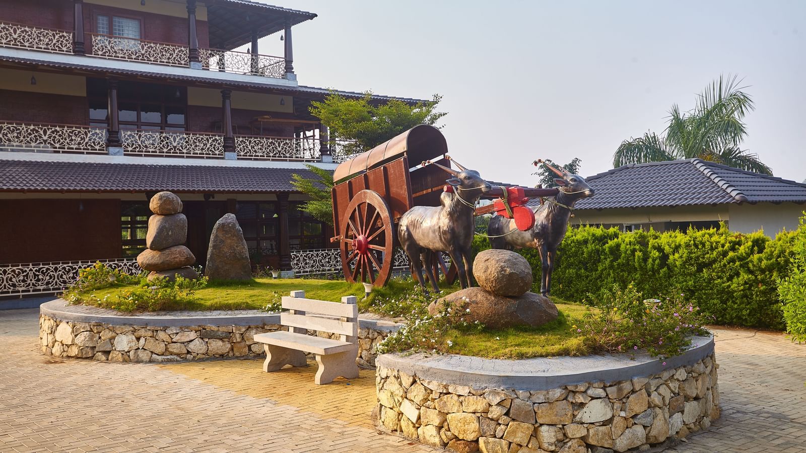 An outdoor seating area with a circular stone-clad planter in the centre, featuring green topiary and benches, set against the backdrop of a multi-storey building - Vintage Trails by Ilara