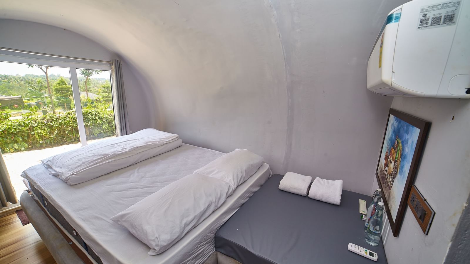 The interior of a modern, capsule-style accommodation unit featuring two single beds with white mattresses and a small bedside table, with a window offering a view of greenery. - Vintage Trails by Ilara