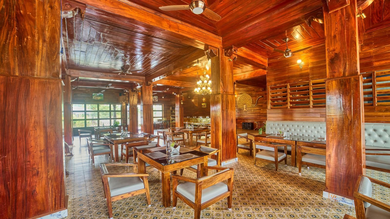 A spacious, warmly lit dining or common area with polished wooden floors, intricate wooden ceilings, and numerous tables and chairs, showing more of the window view. - Vintage Trails by Ilara