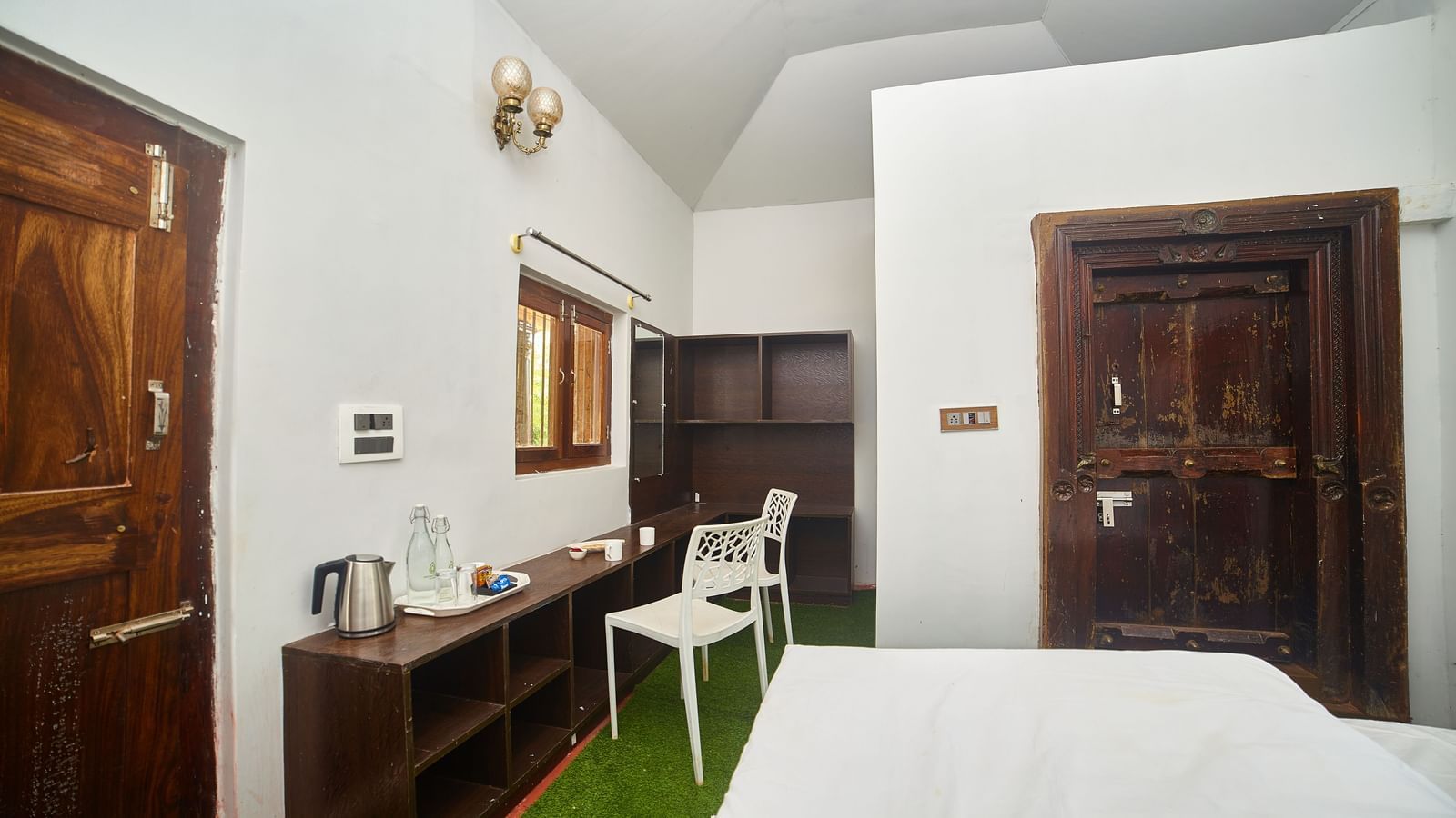 A bright hotel room featuring a bed with white linens, wooden furniture including a cabinet and a white chair, and two dark wooden doors. - Vintage Trails by Ilara