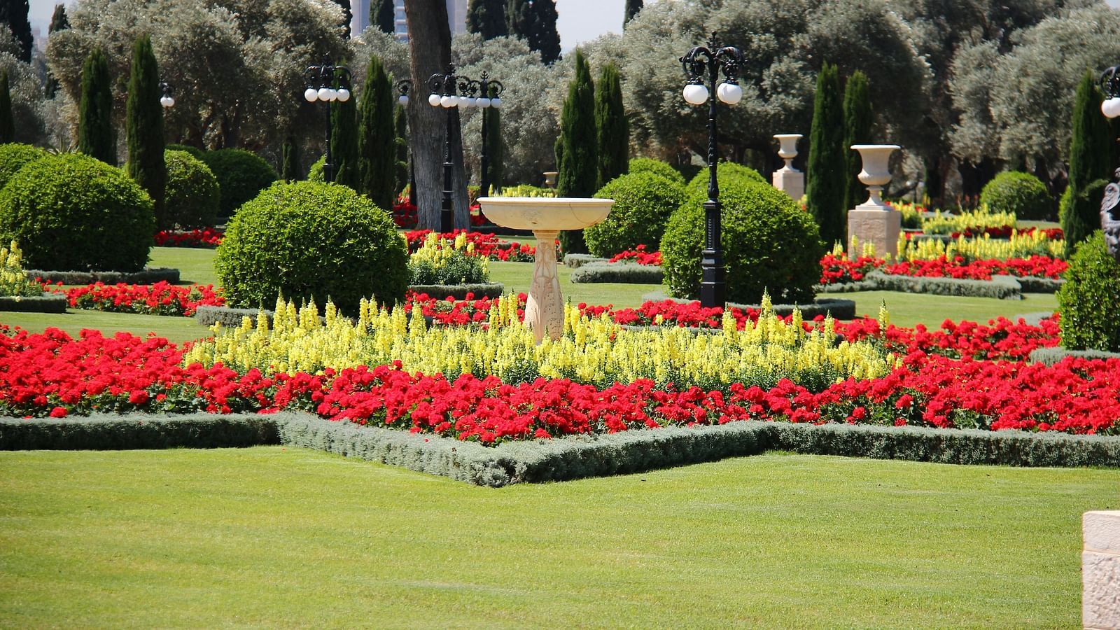 Well-manicured garden with vibrant red and yellow flowers, trimmed bushes, and a central decorative fountain, under a clear blue sky