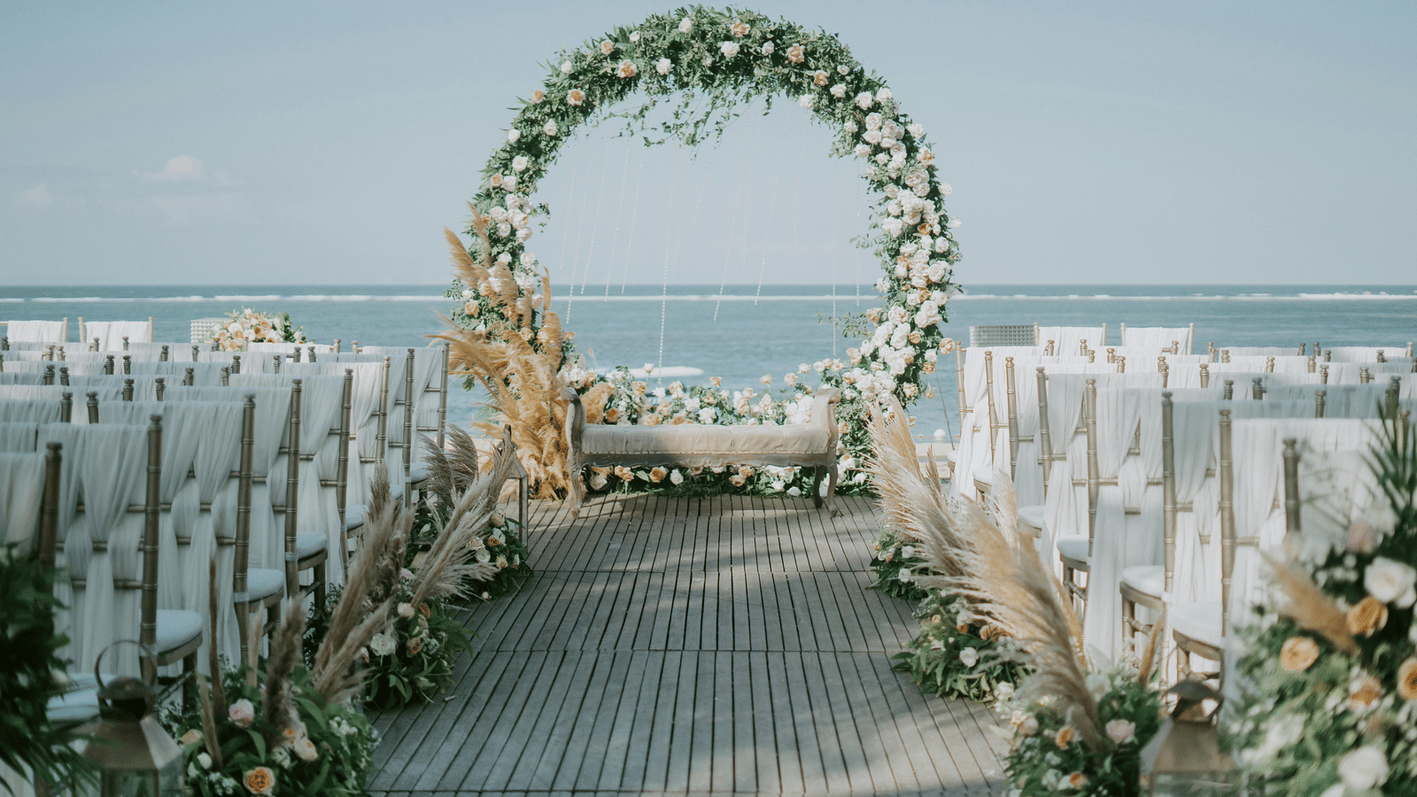 An aisle leading to a floral circular arch, with neatly arranged chairs and the sea in the background.