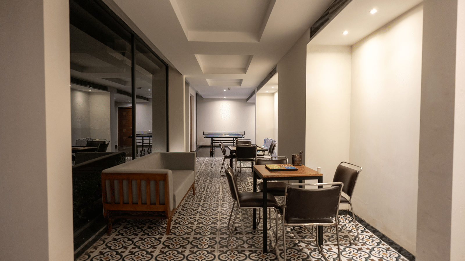 A modern lobby-style acitivity room at Wyndham Garden featuring a patterned tile floor, a long wooden sofa on the left, small dining tables with chairs on the right, ceiling lighting, plain walls and columns, and a large reflective glass wall.