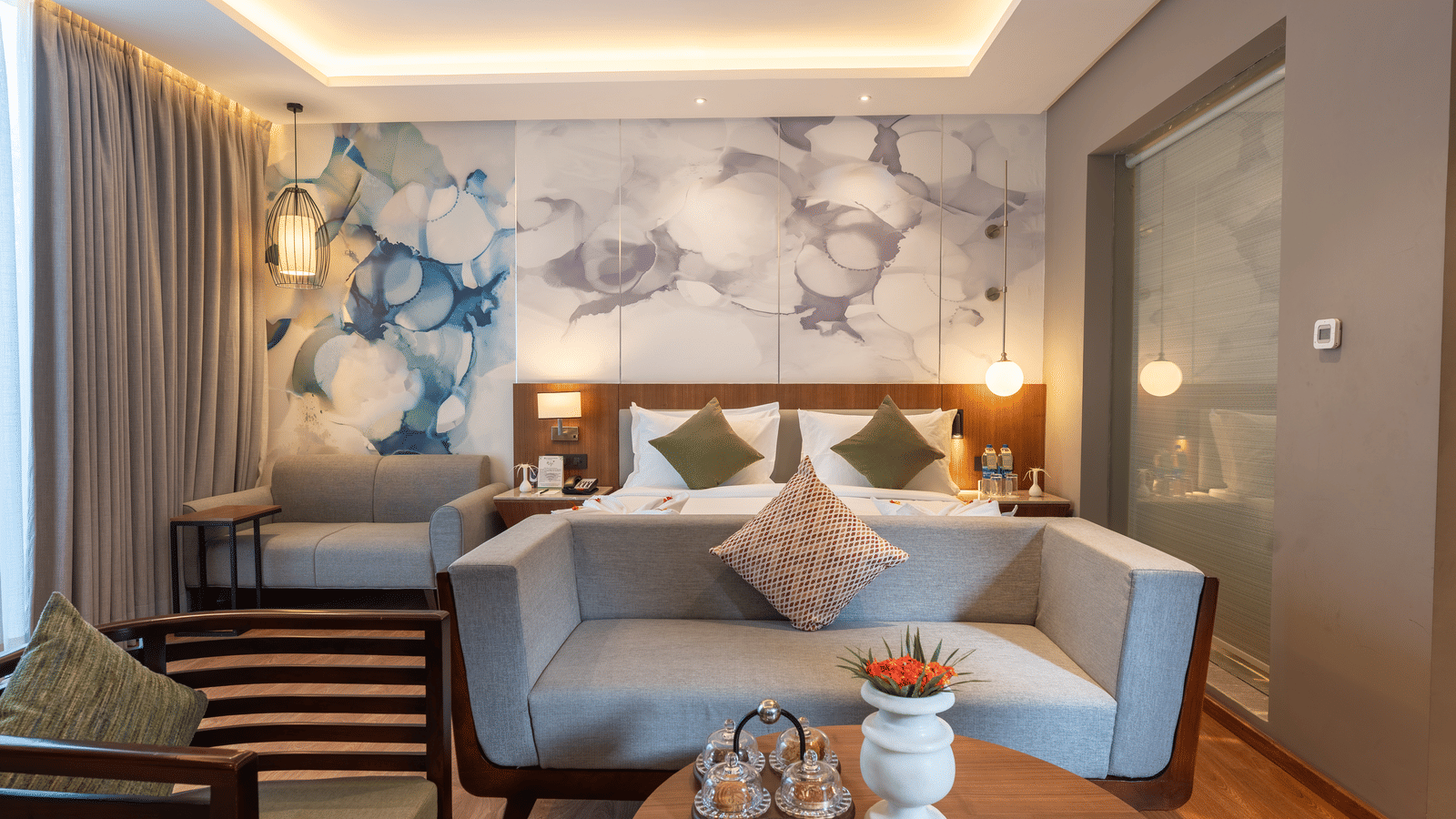 A bedroom at Wyndham Garden with a large beds, a round wooden central table, and a seating area with a wooden armchair visible on the left, warm lighting and heavy curtains covering the window.