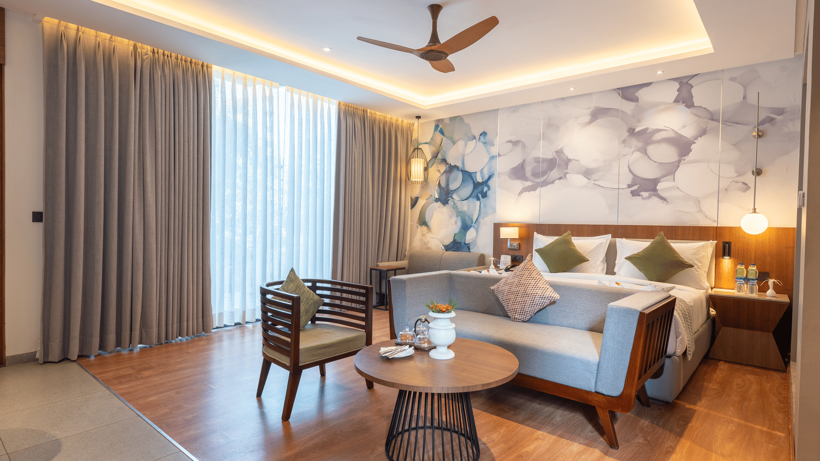 A bedroom at Wyndham Garden with a double bed, wooden armchairs, marble and abstract accent wall, and glass windows draped with long curtain.