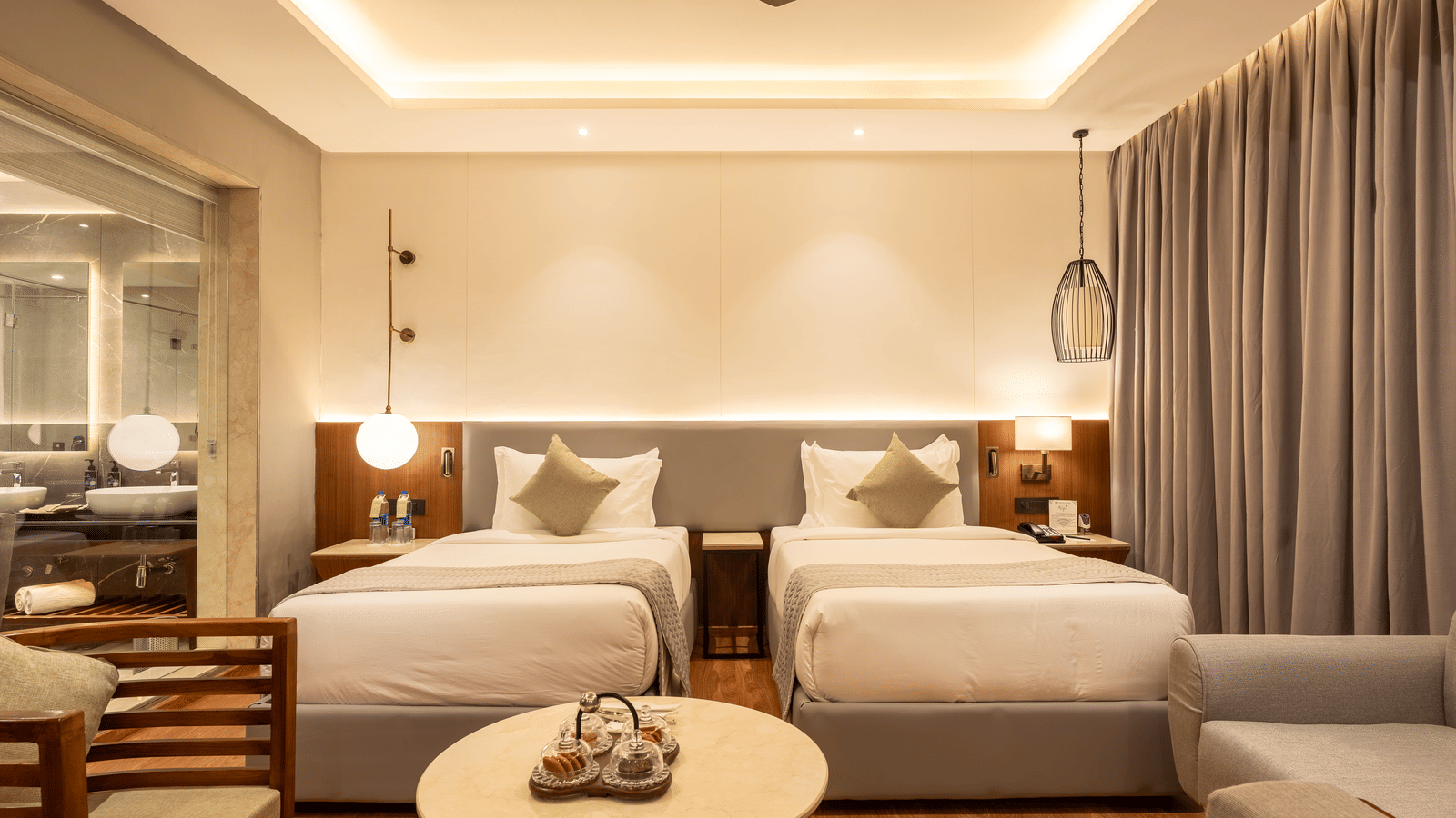 A close-up of twin beds in a guest room at Wyndham Garden showcasing a central bedside table, hanging lighting fixtures,  glass windows draped with long curtains, and wooden furnishing.