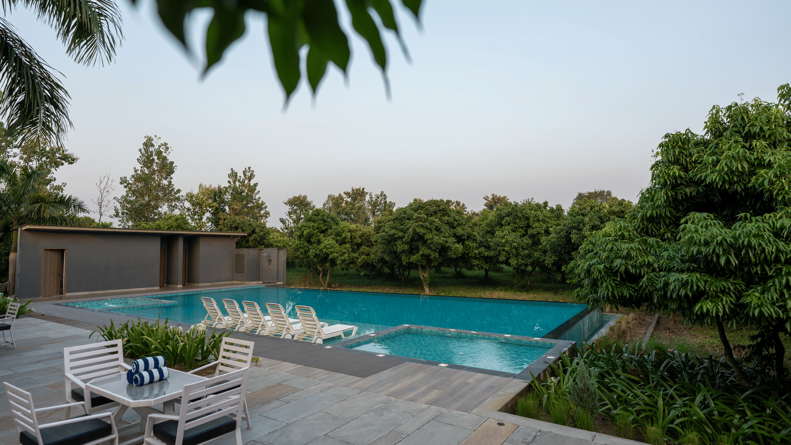 An outdoor seating area at Wyndham Garden with a table and chairs on a stone patio overlooking a large swimming pool, surrounded by lush plants and trees.