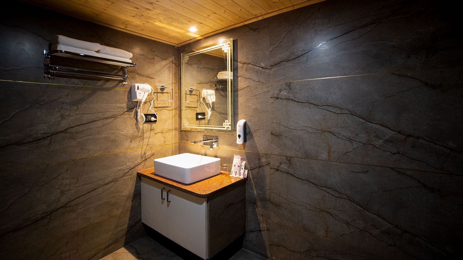Modern bathroom with dark marble-patterned walls and a wooden ceiling, featuring a countertop sink with a mirror above it, wall-mounted hair dryer, soap dispensers, and soft warm lighting - Yashshree Lachen.