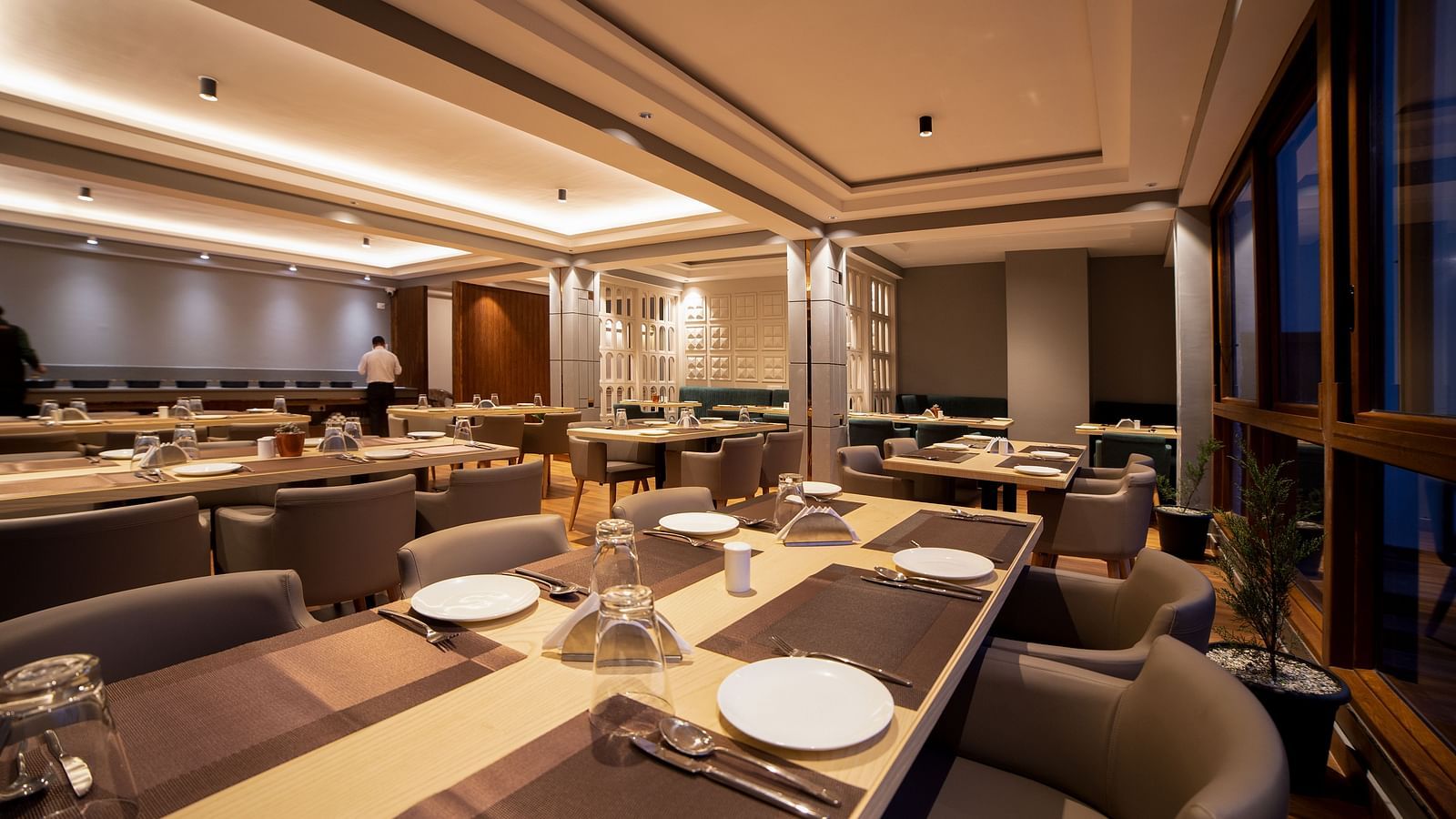 Elegant restaurant interior featuring long tables set with plates, glasses, and silverware, cosy cushioned chairs, warm ambient lighting, and large windows overlooking a nighttime view - Yashshree Pelling Chekhim Retreat.