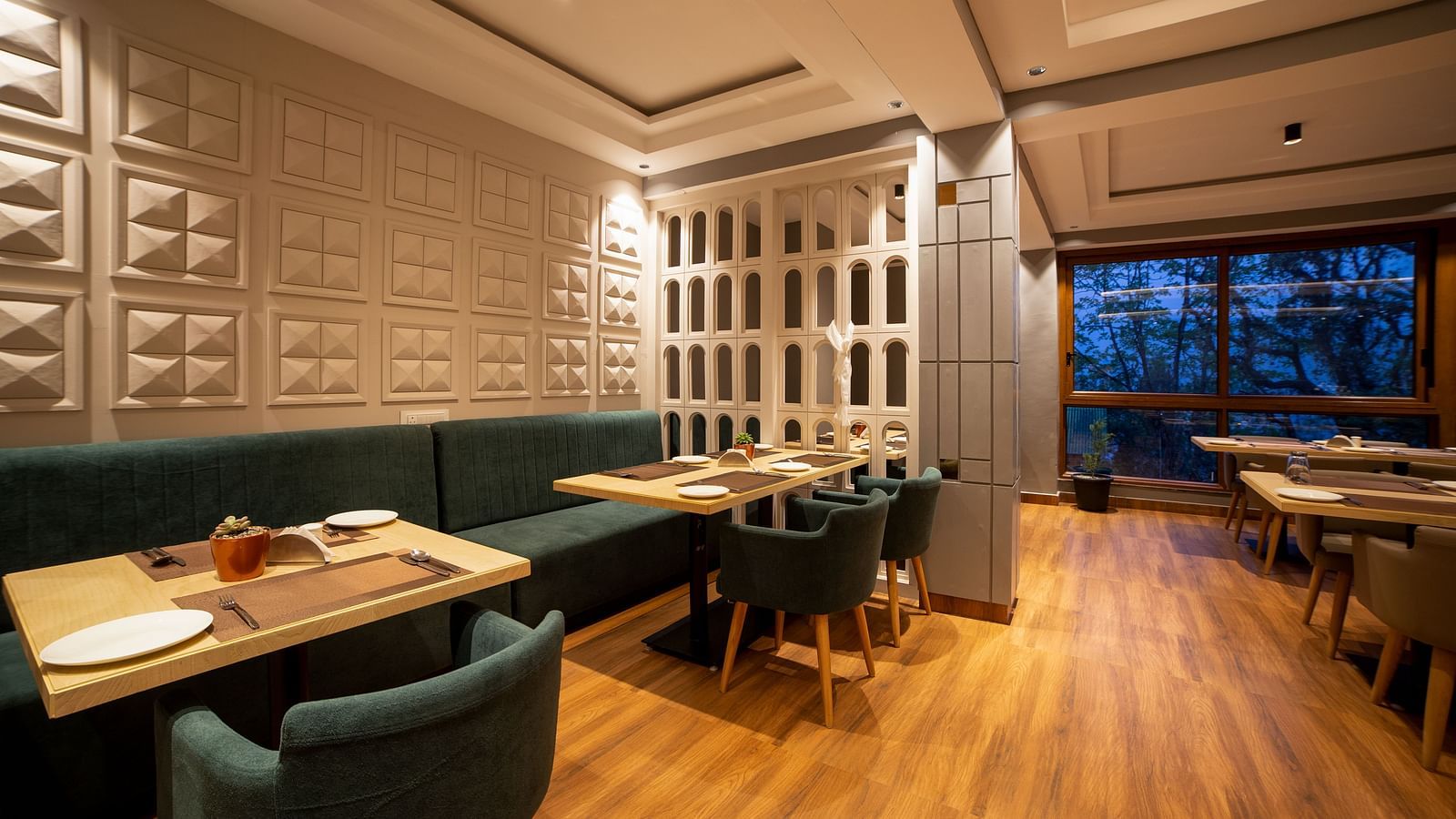 Dining area with wooden flooring, tables and cushioned chairs, a long green upholstered bench along the wall, decorative wall panels, recessed ceiling lighting, and large windows showing trees outside - Yashshree Pelling Chekhim Retreat.