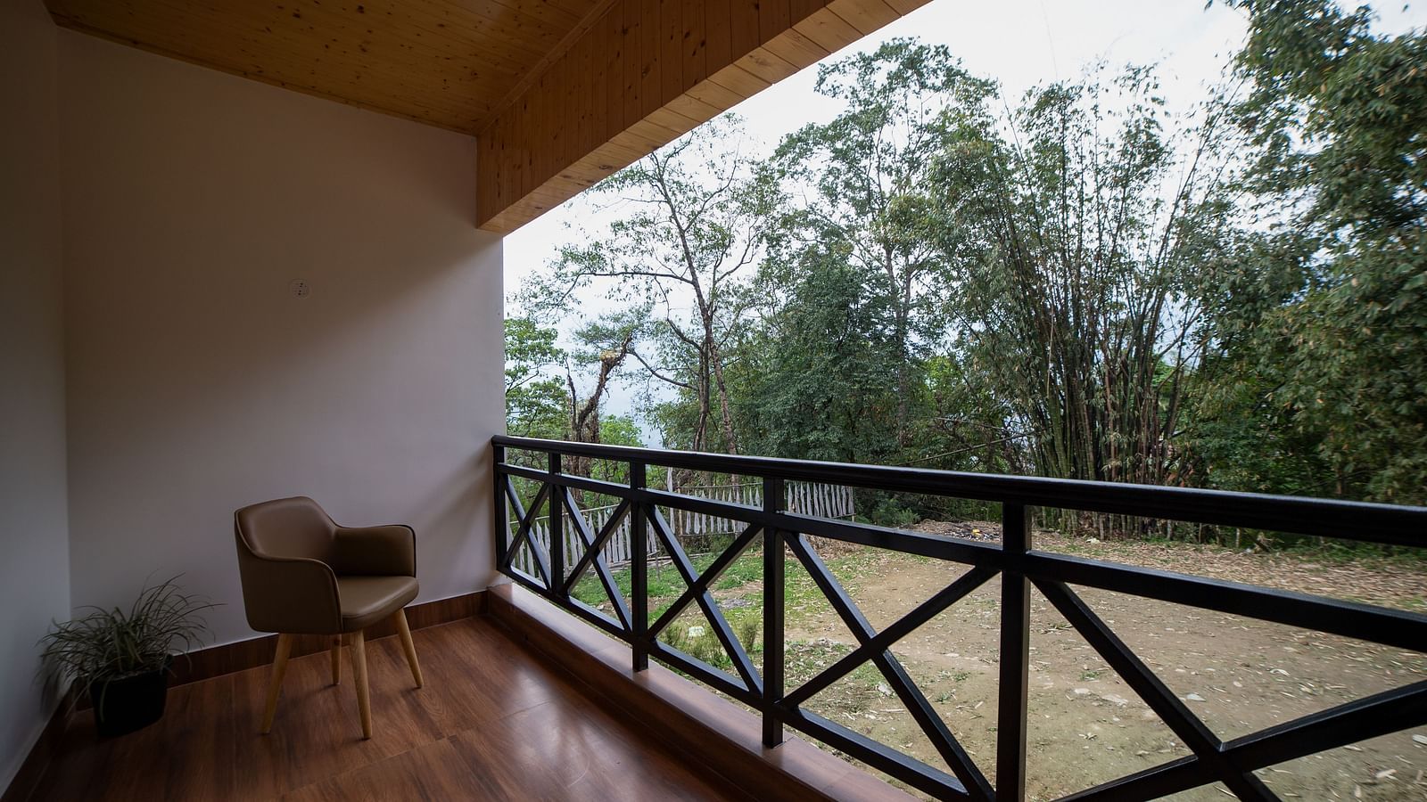 Covered balcony with wooden flooring, a single chair, potted plant, and black railing, overlooking a garden area with trees - Yashshree Pelling Chekhim Retreat.