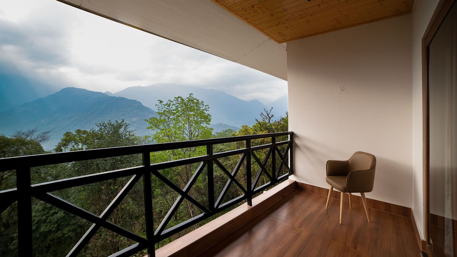 Private hotel balcony with wooden flooring, a single chair, black railing, and a scenic view of lush greenery and misty mountains under a cloudy sky - Yashshree Pelling Chekhim Retreat.