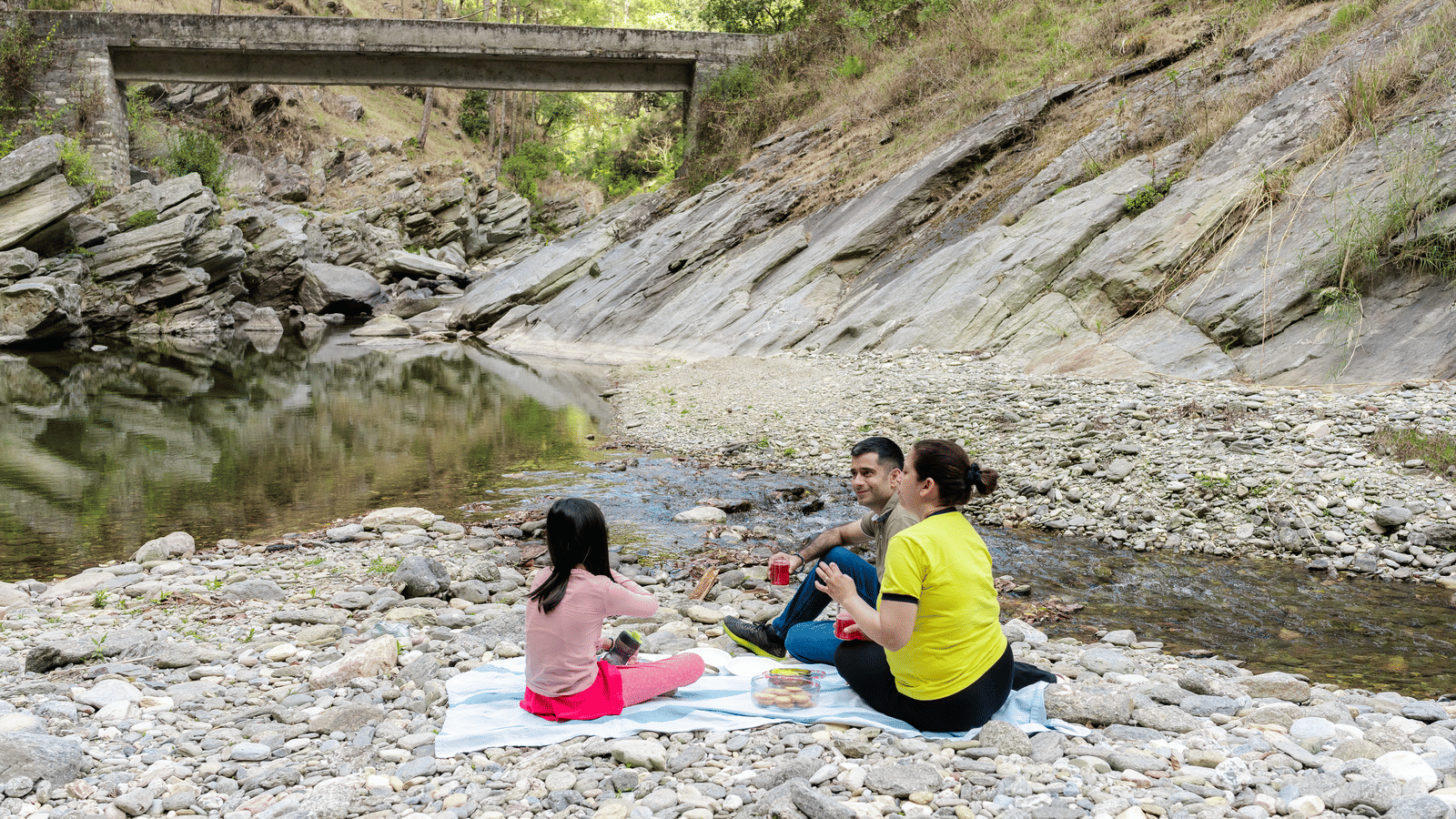 A woman in a yellow shirt and a child in a pink shirt are sitting on a blanket near a river at Ziran Retreat.