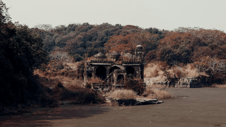Decaying stone ruins with arched windows sit on the edge of a murky river, surrounded by autumn-coloured woodland.