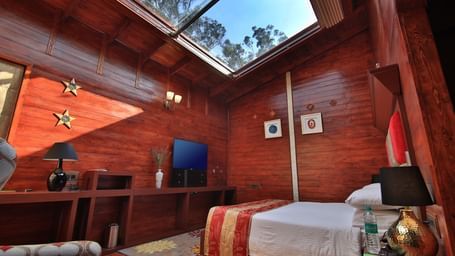 A wide angle image of a room in Ooty featuring a bed, a television and a glass roof flooding the space with sun light - Accord Highland Ooty
