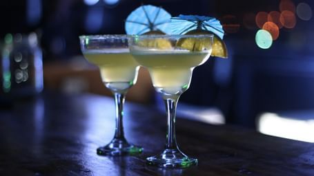 Two margaritas in stemmed glasses with lime wedges and paper umbrellas, on a dark bar counter.