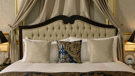A luxurious hotel room features a large bed with a canopy and elegant bedding, flanked by bedside tables with lamps.