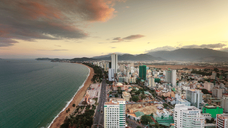 High-rise city buildings line a sweeping sandy coastline beside deep turquoise waters at twilight.