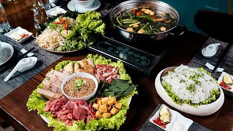 A table set with a variety of dishes including sliced meats, vegetables, noodles, and a pot of broth, ready for a meal.