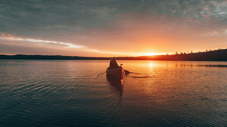 A person kayaking towards land in the far distance with trees and and hills on it and the sun setting in the background