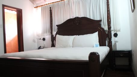 A cozy bedroom is furnished with a dark wooden bed frame, white bedding, and curtains framing a window | Superior Suite | Altamont Court, Jamaica
