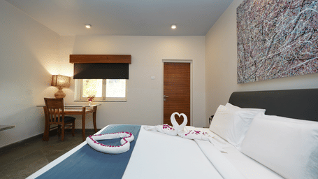 Bedroom with a neatly made bed, a wooden desk with a lamp by the window, and a closed wooden door in the background at Argo By Trance, Goa
