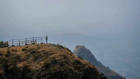 A hazy, wide landscape shot from a dry hilltop viewpoint with a small wooden fence, overlooking layered mountains fading into the distance.