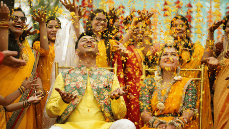 A joyful haldi ceremony scene with a couple in traditional yellow attire being showered in flower petals by smiling family and friends.
