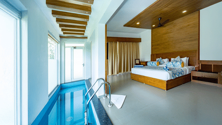 A wide-angle shot showcases a modern, spacious bedroom with a king size bed dressed in white linens with a blue floral bed runner at Aveda Mountains and Mist Resort and Spa, Munnar.
