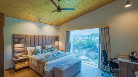 A room at Bamboo Saa Resort & Spa, Udaipur, featuring a large bed, bedspreads, pillows, a fan. a table, and windows with curtains.