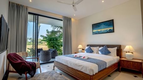 A room with balcony, spacious bed with bedside tables and lamps, artwork above the headboard, and outdoor view through glass doors