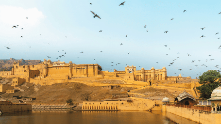 A far-out view of Amber Fort with a water body in the foreground and birds flying in the sky. A must-visit while exploring activities in Jaipur.
