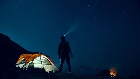 A person standing near a glowing tent, gazing at the night sky
