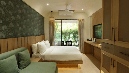 A bedroom features a sofa, a king size bed, a television unit, and large glass doors leading out to a garden terrace at Bougainvillea, Alibaug.