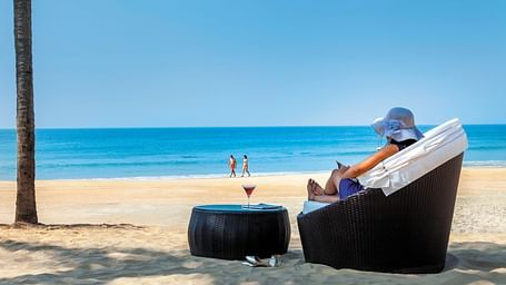 a woman sitting on the chair with a drink on the table on the beach - Caravela Beach Resort Goa.