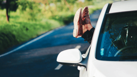 A person's feet are sticking out of the passenger side window of a white motorcar travelling along a country road.