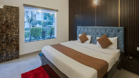 The bedroom of the Family Cottage at Corbett Holiday Forest Resort, Ramnagar, featuring a double bed with a tufted head board and a window beside it.
