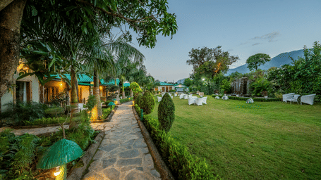 Day-time view of a lush green lawn and pathway leading to the cottage-style accommodations at Corbett Nirvana Resort.