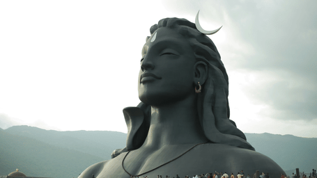 A far out view of the 112 foot Adiyogi Temple inside Coimbatore Isha Temple with numerous people walking at the ground level and mountains in the background.