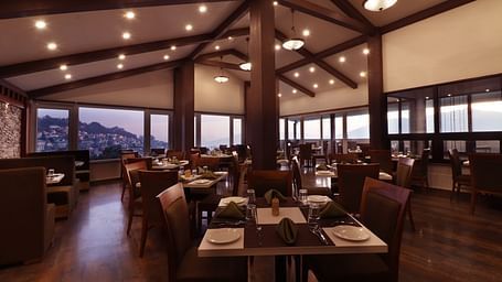 A modern styled rooftop dining space featuring wooden furniture and arranged table set with tableware and folded napkins and huge windows that opens to hilly cityscape at the backdrop.