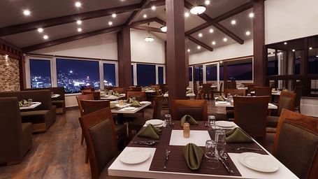 A close up view of the rooftop dining hall at Denzong Shangrila Lords Inn Gangtok featuring wooden furnishing, arranged table set with tableware and large windows opening to the evening view of illuminated town.