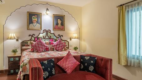 A bedroom with a patterned bedspread, colourful cushions, a red sofa, and artwork in arched niches | Dera Rawatsar, Jaipur