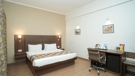 A double bedroom at Diamond Hotel Varanasi features a wooden desk, a mini-fridge, and framed wall art.