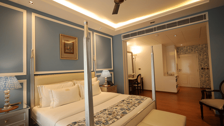 A bedroom with a four-poster bed, blue panelled walls, and a recessed ceiling with warm lighting, leading into an adjacent room with a desk and chair | Dileep Kothi, Jaipur