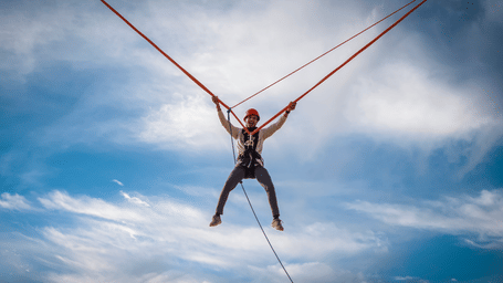 Man wearing a red helmet launched into the air attached to elastic ropes outdoors under clear sky.