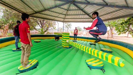 Man jumping over a rotating arm on a green and yellow inflatable wipeout game under a roof structure.