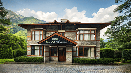 Front facade of stone and wooden resort building with mountain peaks in background at DLS Dev Lok, Manali.   