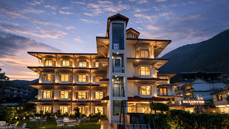 Exterior view of DLS Meghavan Resort, Dharamshala, a multi-story hotel building with glowing windows, a central glass elevator shaft, an outdoor lawn with dining tables and a swing bench, framed by mountains.